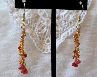 May include: A pair of dangle earrings with a gold chain and red and orange glass beads. The earrings have a flower-shaped charm at the bottom.