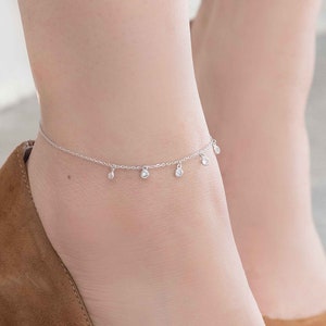 May include: A silver anklet with dangling, clear gemstones. The anklet is worn with brown suede high heels. The shoes have pointed toes and stiletto heels. The background is a light, neutral color.