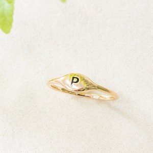 May include: A gold signet ring with the letter "P" engraved on the top. The ring is on a white surface.