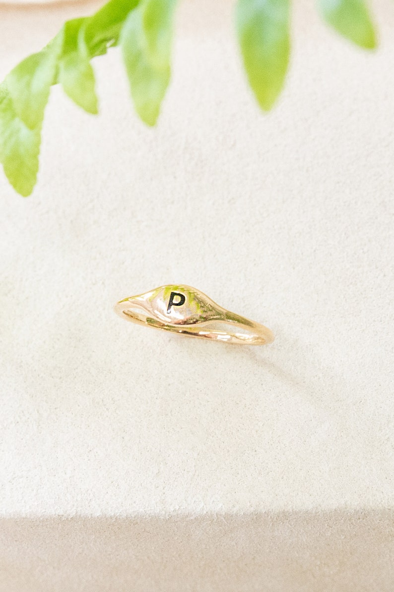 May include: A gold signet ring with the letter "P" engraved on the top. The ring is on a white surface.