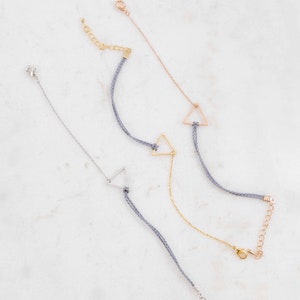 May include: Three silver, gold, and rose gold triangle charm bracelets with gray string and lobster claw clasps.