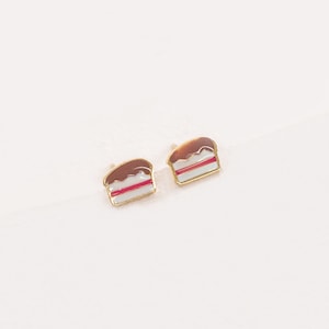 May include: A pair of gold-tone earrings with a small cake design. The cake is white with a pink layer and brown frosting.