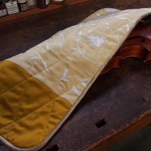 May include: A quilted violin case with a floral pattern on the top and a solid mustard yellow colour on the bottom. The case is made of a soft fabric and has a zipper closure.