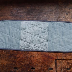 Op de afbeelding: Een rechthoekige, gewatteerde placemat in blauw- en grijstinten. Het middengedeelte heeft een bloemenpatroon, terwijl de omliggende gebieden een diamantgestikt ontwerp hebben. De randen zijn afgewerkt met een donkergrijze bies.