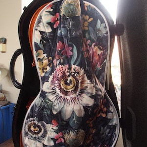 May include: A cello case with a floral quilted interior. The interior fabric features a dark background with large, colorful flowers in shades of pink, purple, yellow, and white. The case is open, revealing the instrument's neck and strings.