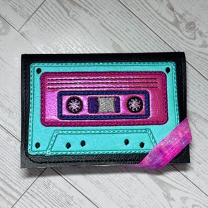May include: A black wallet with a teal and pink cassette tape design. The cassette tape has a metallic sheen and is outlined in black. A pink and purple ribbon is attached to the wallet.
