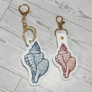 May include: Two seashell keychains. One keychain features a blue embroidered seashell on a white leather backing, attached to a gold-colored clasp and ring. The other keychain has a red embroidered seashell on a white leather backing, with a gold-colored clasp.