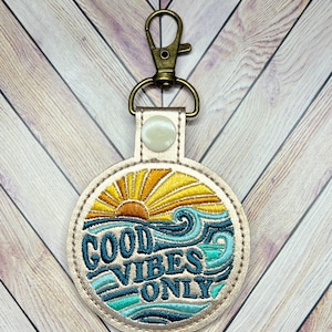May include: A gold-colored leather keychain with a round, embroidered patch that says "Good Vibes Only". The patch features a blue and green wave design with a yellow sun rising above the waves.