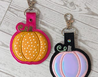 May include: Two pumpkin-shaped keychains. One is orange with white polka dots and a pink border. The other is a light purple with a pink border and a black outline. Both have silver clasps and green vine accents.