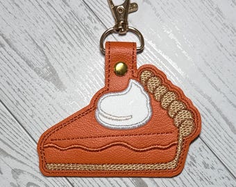 May include: An orange pumpkin pie slice keychain made of textured material with a white embroidered dollop of whipped cream on top, a gold-colored metal lobster clasp, and a gold snap button closure on the orange leather-like tab attachment.