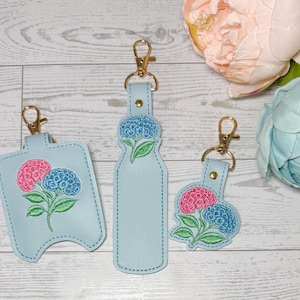 May include: Three light blue keychains with gold-colored hardware. Each keychain features an embroidered hydrangea design in pink and blue. The keychains are in different shapes, including a bottle shape and a hand sanitizer holder.