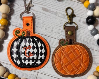 May include: Two pumpkin-shaped keychains. One is orange with a black and white argyle pattern and the letter "C". The other is orange with a quilted design. Both have metal clasps and brown or orange leather straps.