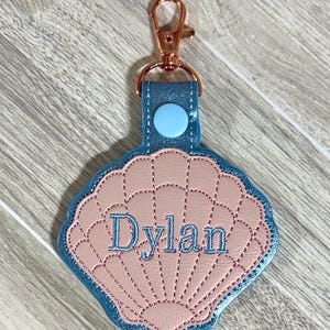 May include: A seashell-shaped keychain with the name "Dylan" embroidered in blue thread. The keychain features a light pink shell design with a blue border and a blue strap with a button. It has a gold-colored clasp.