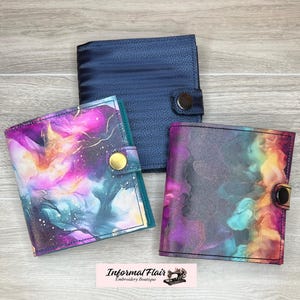May include: Three different fabric covered notebooks with a button closure. The notebooks are in a galaxy print, a blue print, and a purple and teal print.