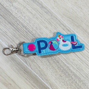 May include: A light blue keychain with the word "POOL" embroidered in dark blue. The keychain features a pink dot, a beach ball, and a white unicorn with a pink and yellow mane. A silver clasp is attached.