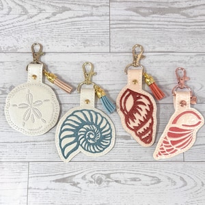May include: Four seashell-themed keychains with gold-colored clasps and small tassels. The keychains feature a sand dollar, a nautilus shell, a red shell, and a pink shell design, all on a cream-colored background.