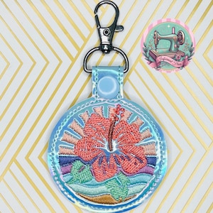 May include: A light blue round keychain with a silver metal clasp. The keychain features an embroidered design of a pink hibiscus flower with green leaves and blue waves. The background of the keychain is iridescent.