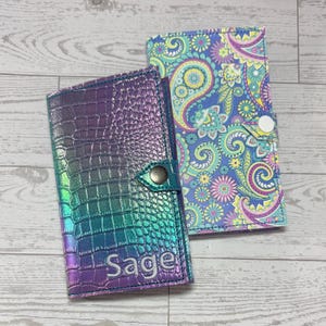 May include: Two decorative passport holders. One is iridescent with a crocodile skin texture and the name "Sage" embroidered on the front. The other has a paisley pattern in shades of blue, purple, and yellow. Both have a snap closure.