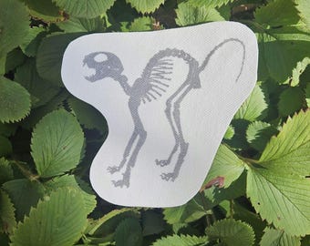 Könnte beinhalten: Ein graues Katzenskelett-Design, das auf einem weißen, unregelmäßig geformten Aufnäher gedruckt ist. Das Katzenskelett ist in einer stehenden Position mit gekrümmtem Schwanz dargestellt. Der Aufnäher befindet sich vor einem Hintergrund aus grünen Blättern.