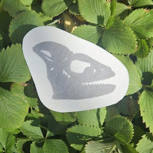 May include: A white fabric patch with a grey silhouette of a dinosaur skull.