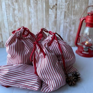 May include: Five red and white striped fabric gift bags with red drawstrings. The bags are arranged in a pile on a white surface.