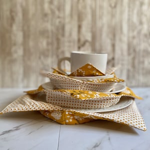 May include: A set of yellow and white fabric pot holders with a floral pattern and polka dots. The pot holders are stacked on top of each other and a white mug is sitting on top of the stack.