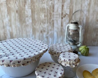 May include: Set of four reusable food covers in a beige and pink floral pattern. The covers are made of fabric and have a ruffled edge. They are shown covering bowls and a jar.