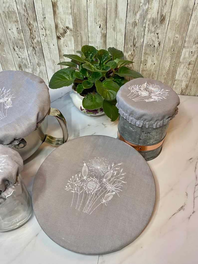 May include: A collection of gray fabric covers with white embroidered floral designs. The covers are placed on various containers, including a pot, a jar, and a metal bucket. A potted plant with green leaves is in the background.
