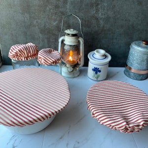 May include: Red and white striped fabric covers for bowls and jars. The covers are made of a cotton fabric with a red and white stripe pattern. The covers are shown on a white surface with a vintage lantern and other decorative items.