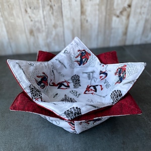 May include: A red and white fabric bowl with a starburst shape. The bowl is lined with a white fabric featuring a repeated pattern of Spiderman swinging from buildings. The bowl is sitting on a gray surface.