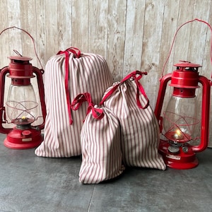 May include: Red and white striped fabric drawstring bags in various sizes with red ribbon ties. Two red vintage lanterns with glass chimneys flank the bags. The lanterns have a handle and a fuel gauge.