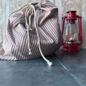May include: A red and white striped drawstring bag with a cream-colored rope closure. A red lantern with a glass enclosure sits in the background. The bag is made of fabric and is gathered at the top.