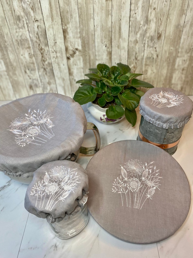 May include: A collection of gray fabric bowl covers with white embroidered floral designs. The covers are in various sizes, fitting over a bowl, a jar, and a metal container. A potted plant is in the background.