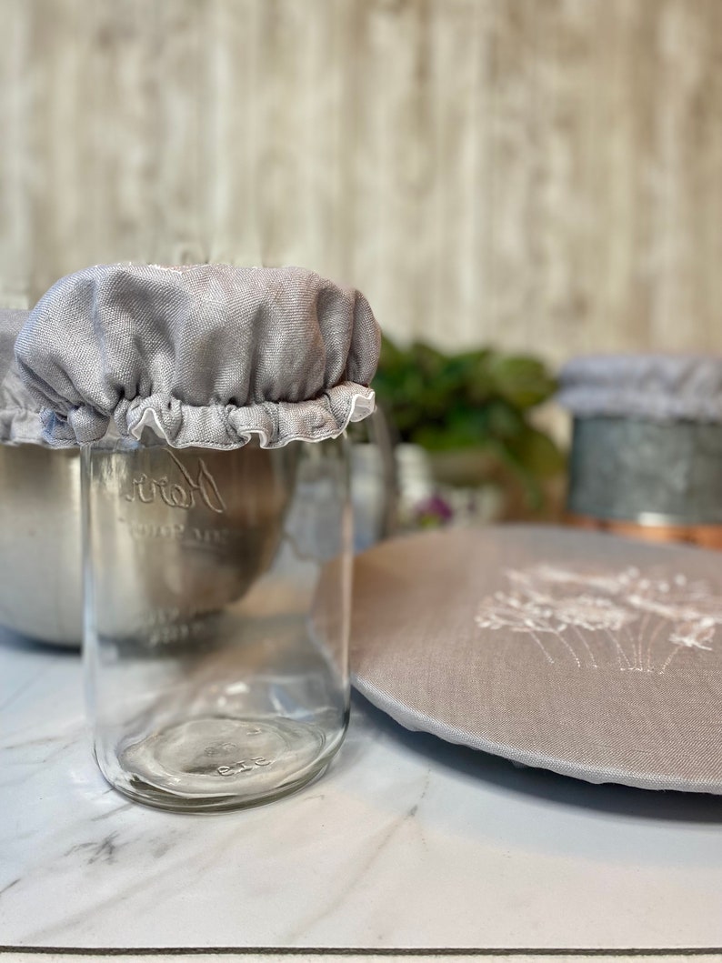 May include: Clear glass jar with a gray fabric lid and white ruffled trim. A matching gray fabric lid with white floral embroidery is also visible. The jar is on a white marble surface.