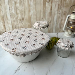 May include: Set of three reusable food covers with a white and brown gingham pattern and a chicken print. The covers are made of fabric and have a ruffled edge. They are shown covering a bowl and two glass jars.