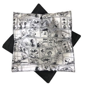 May include: A white and black fabric coaster with a Peanuts comic strip design. The coaster features various Peanuts characters, including Snoopy, Charlie Brown, and Lucy. The text "HE SAY THAT ONCE YOU'RE OVER THE HILL, YOU BEGIN TO PICK UP SPEED" is printed on the coaster.