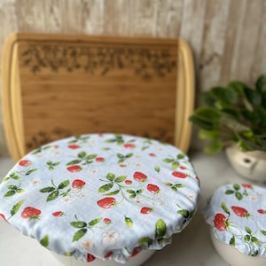 May include: Two white bowls covered with light blue fabric lids featuring a strawberry pattern. The lids have red strawberries, green leaves, and white flowers. A wooden cutting board is in the background.