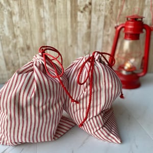 May include: Two red and white striped fabric gift bags with red drawstrings. The bags are sitting on a white surface.