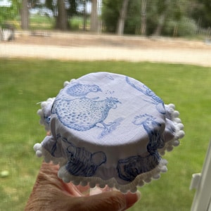 May include: A white and blue fabric jar cover with a chicken illustration. The cover has a ruffled edge with white pom-poms. The jar cover is held in front of a window with a green background.