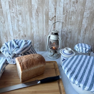 May include: A loaf of white bread on a wooden cutting board with a bread knife. There are three blue and white striped fabric covers on bowls and a vintage oil lamp.