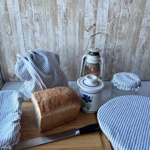 May include: A loaf of bread sits on a wooden cutting board with a knife, a blue and white ceramic jar, and three blue and white striped fabric covers.