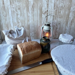 May include: A loaf of bread on a wooden cutting board with a knife, a ceramic jar, and four cloth covers with a striped pattern. The covers are used to store food in the refrigerator.