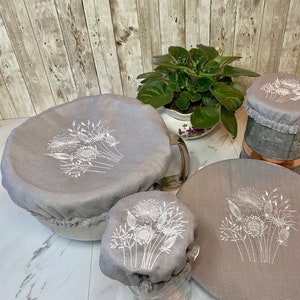 May include: A collection of gray fabric bowl covers with white embroidered floral designs. The covers are in various sizes, designed to fit different bowls and containers. The covers are displayed on a white surface with a wooden background.