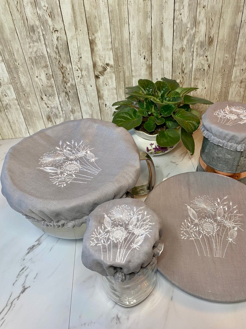 May include: A collection of gray fabric bowl covers with white embroidered floral designs. The covers are in various sizes, designed to fit different bowls and containers. The covers are displayed on a white surface with a wooden background.