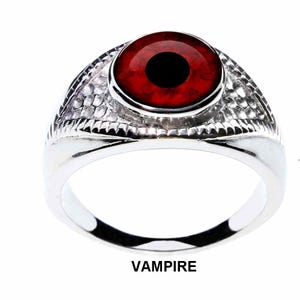 Może przedstawiać: Srebrny pierścionek z dużym, okrągłym, czerwonym kamieniem z czarnym środkiem, przypominającym oko. Pierścionek ma teksturowaną obręcz, a pod nim wydrukowano słowo "VAMPIRE".