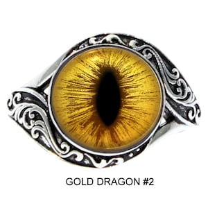 Może przedstawiać: Srebrny pierścień z dużym, złoto-żółtym okiem smoka z czarną źrenicą. Pierścień ma ozdobny, wirujący wzór wokół oka i tekst "GOLD DRAGON #2" na dole.