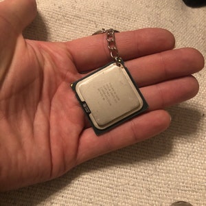 May include: A silver keychain with a small, square, silver CPU chip attached to it. The chip has text on it that reads "Intel Core i7 9700K 8-Core 3.6 GHz 12MB Cache 95W TDP".