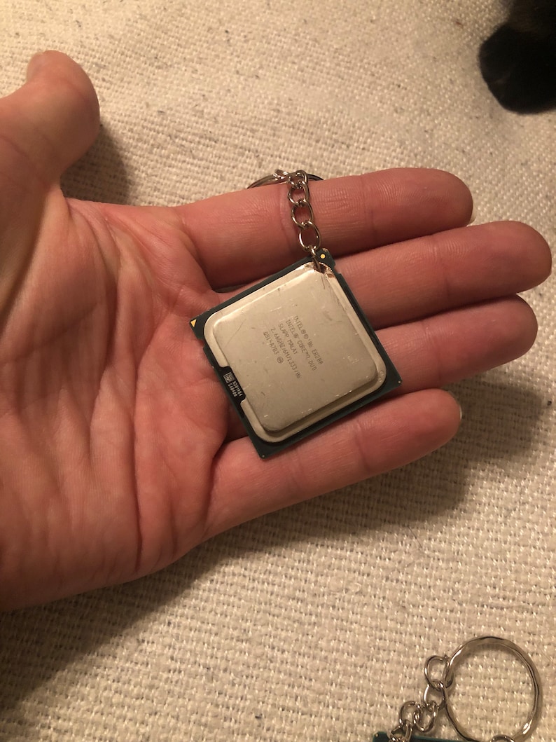 May include: A silver keychain with a small, square, silver CPU chip attached to it. The chip has text on it that reads "Intel Core i7 9700K 8-Core 3.6 GHz 12MB Cache 95W TDP".