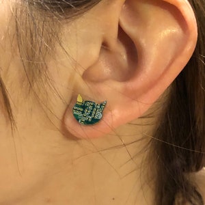 May include: A close-up of an ear with a cat-shaped stud earring. The earring is green with a circuit board design and a yellow accent on the ear. The earring is a unique and quirky accessory.