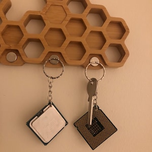 May include: A wooden honeycomb-shaped key holder with two keychains attached. One keychain has a silver key and a gold-colored CPU chip. The other keychain has a silver ring and a white CPU chip.